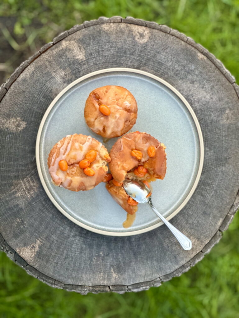 Three glazed sea buckthorn muffins on a grey plate, topped with bright orange berries, served on a rustic wooden surface outdoors.