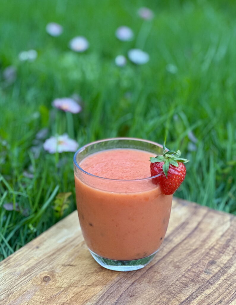 Glass of sea buckthorn smoothie with mango and strawberries on a wooden board, topped with a fresh strawberry, outdoors on green grass.